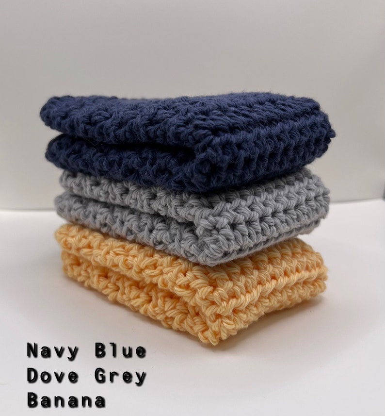 May include: A stack of three crocheted dishcloths in navy blue, dove grey, and banana yellow.