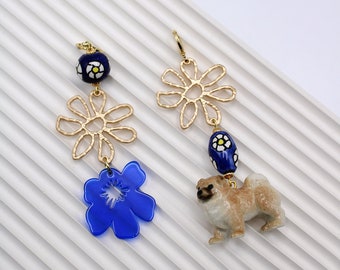 以下が含まれることがあります： 青と白の花柄のアクセントが付いた、ゴールドのドロップイヤリングのペア。片方のイヤリングには青い花のチャーム、もう片方には小さなセラミック製の犬のチャームが付いています。