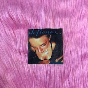 May include: A black and white patch with the band name "Deftones" and the album title "Around the Fur" printed on it. The patch features a photo of a woman with dark hair and a black top.