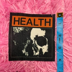May include: A black and white patch featuring a skull graphic. The patch has the word "HEALTH" in orange block letters at the top. A blue measuring tape is next to the patch.
