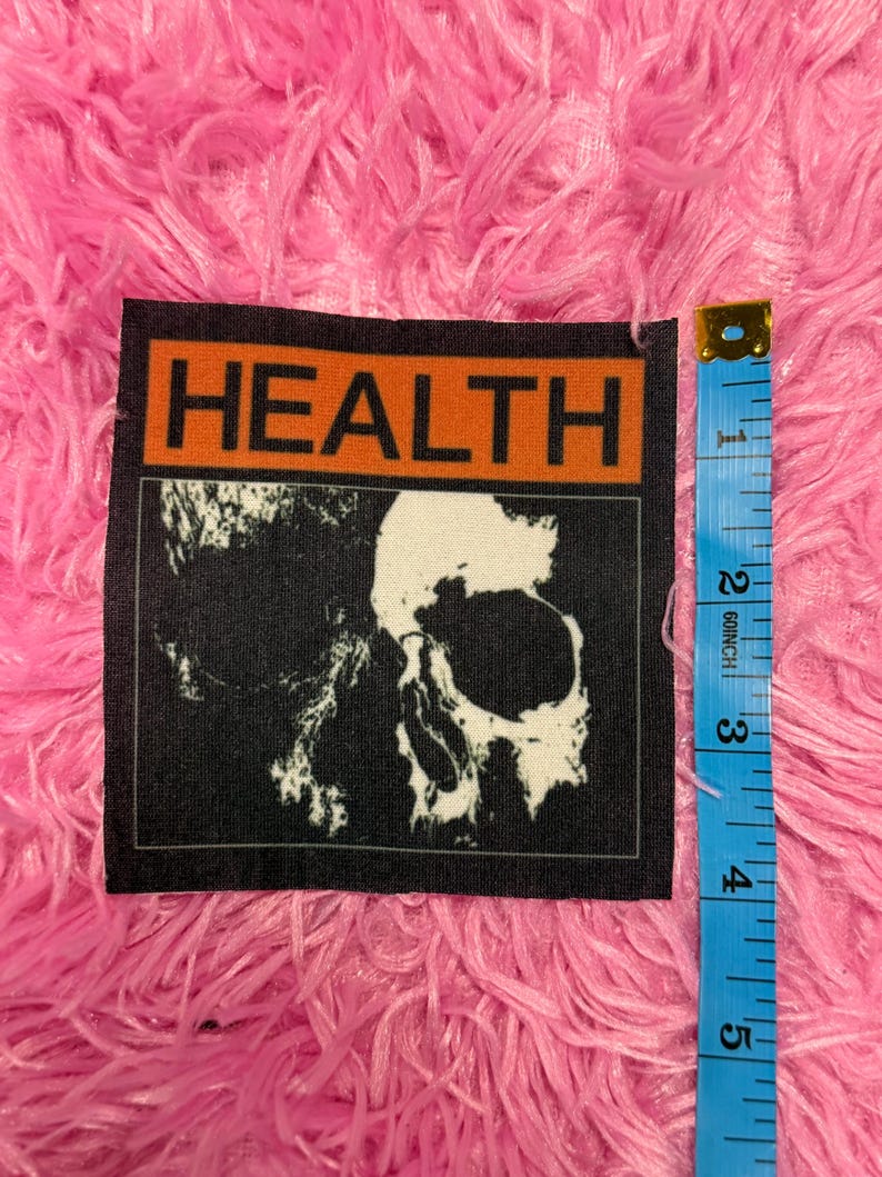 May include: A black and white patch featuring a skull graphic. The patch has the word "HEALTH" in orange block letters at the top. A blue measuring tape is next to the patch.