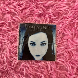 May include: A black and white image of a woman with dark hair and blue eyes. The image is on a black background with the text "Evanescence Fallen" in white.
