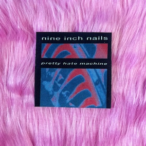 May include: A black square patch with the text "nine inch nails" above "pretty hate machine". The background of the patch features red and blue abstract shapes. The patch is set against a pink, fluffy background.