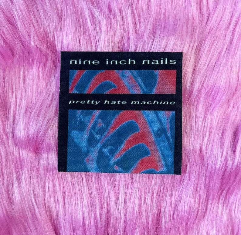 May include: A black square patch with the text "nine inch nails" above "pretty hate machine". The background of the patch features red and blue abstract shapes. The patch is set against a pink, fluffy background.