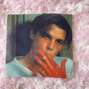 May include: A close-up portrait of a man with dark hair, wearing a white shirt. His right hand is covered in red, as if he has been injured.