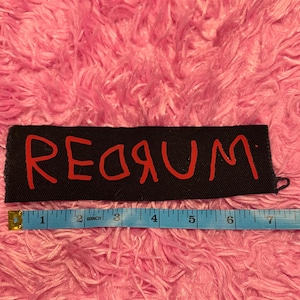 K&ouml;nnte beinhalten: Schwarzer rechteckiger Aufn&auml;her mit dem Wort "REDRUM" in roter Schrift. Unter dem Aufn&auml;her befindet sich ein blaues Ma&szlig;band mit Zollangaben. Der Hintergrund ist eine rosa, flauschige Oberfl&auml;che.