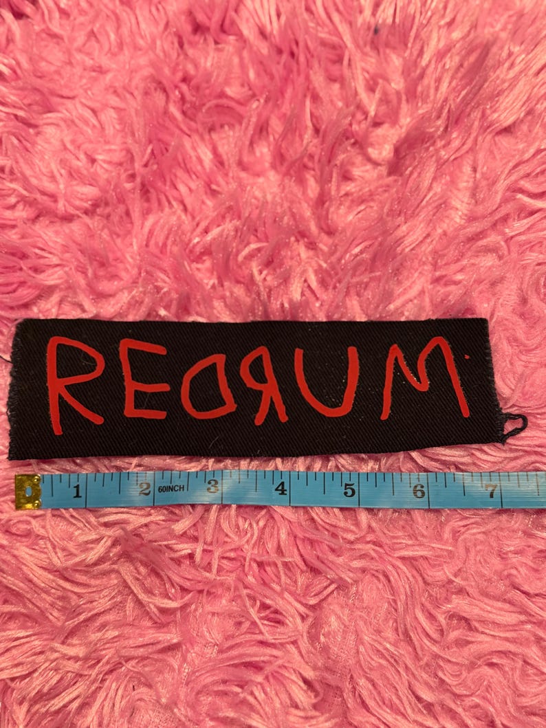 K&ouml;nnte beinhalten: Schwarzer rechteckiger Aufn&auml;her mit dem Wort "REDRUM" in roter Schrift. Unter dem Aufn&auml;her befindet sich ein blaues Ma&szlig;band mit Zollangaben. Der Hintergrund ist eine rosa, flauschige Oberfl&auml;che.