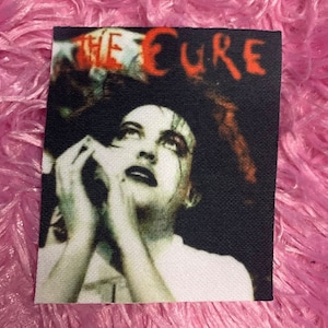 May include: A black and white image of a woman with dark hair and dark eye makeup. The woman is looking up and her hands are clasped together. The text "THE CURE" is written in red letters across the top of the image.