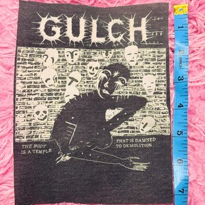 May include: A black and white patch with the word "GULCH" and a graphic of a figure and faces. The text "THE BODY IS A TEMPLE" and "THAT IS DAMNED TO DEMOLITION" are included. The patch is approximately 7.6 cm wide.