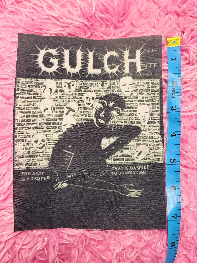 May include: A black and white patch with the word "GULCH" and a graphic of a figure and faces. The text "THE BODY IS A TEMPLE" and "THAT IS DAMNED TO DEMOLITION" are included. The patch is approximately 7.6 cm wide.