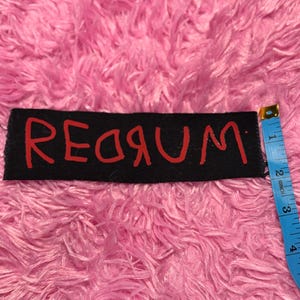 K&ouml;nnte beinhalten: Schwarzer rechteckiger Aufn&auml;her mit dem Wort "REDRUM" in roter Schrift. Der Aufn&auml;her ist etwa 7,6 cm lang. Der Hintergrund ist ein flauschiges, rosa Material.