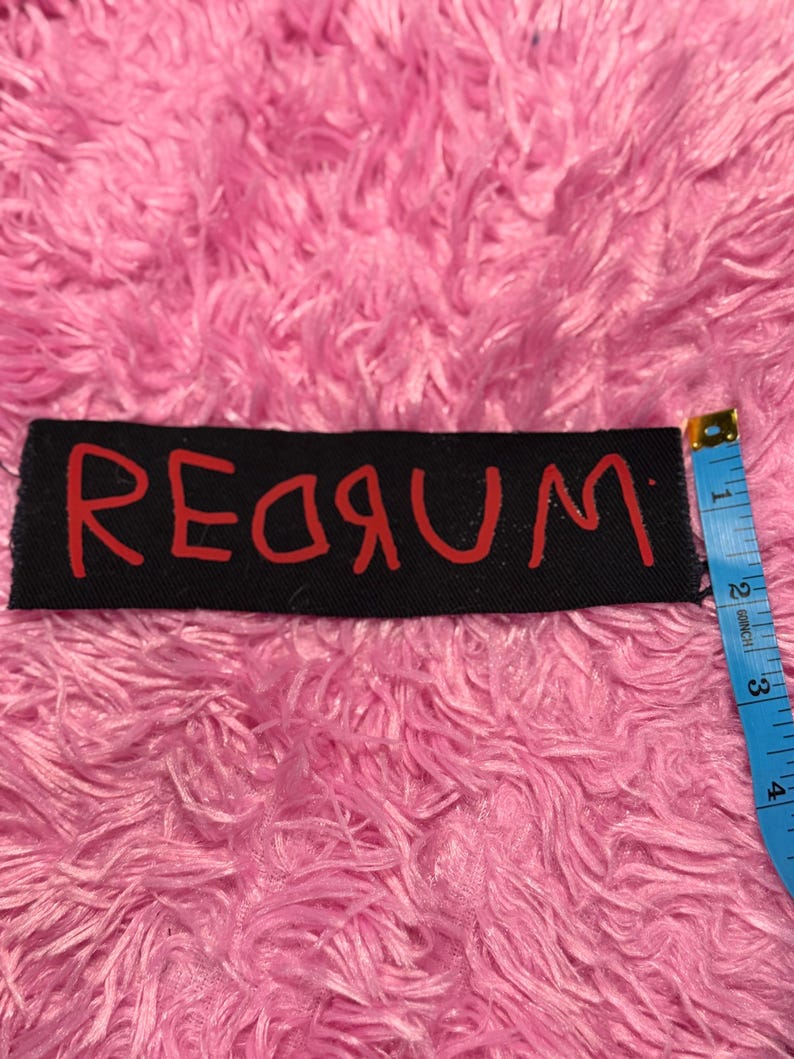 K&ouml;nnte beinhalten: Schwarzer rechteckiger Aufn&auml;her mit dem Wort "REDRUM" in roter Schrift. Der Aufn&auml;her ist etwa 7,6 cm lang. Der Hintergrund ist ein flauschiges, rosa Material.