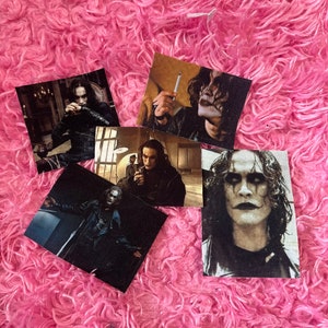 May include: Five black and white photos of a man with dark hair and pale skin wearing a black coat and a black shirt. The man has dark makeup around his eyes. The photos are on a pink fuzzy surface.