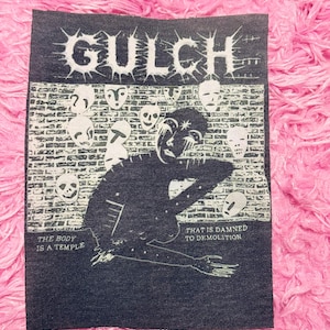 May include: A dark blue patch with the word "GULCH" in white, spiky font. The patch features a black illustration of a figure and skulls, with the phrases "THE BODY IS A TEMPLE" and "THAT IS DAMNED TO DEMOLITION".