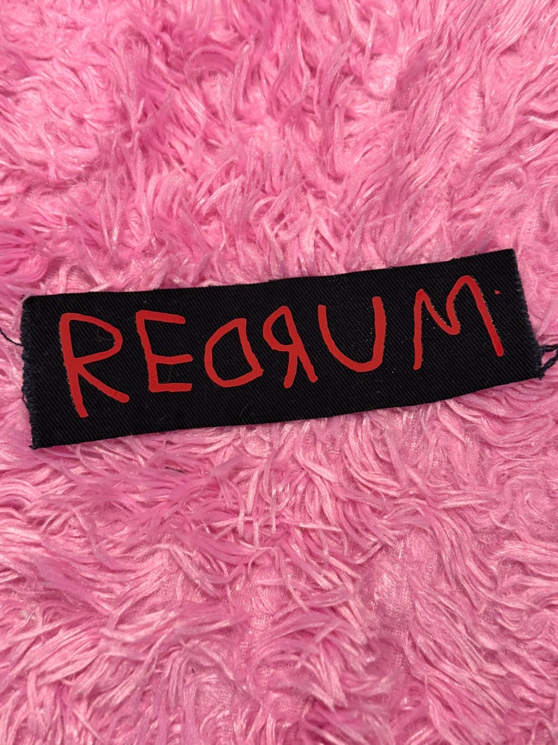 K&ouml;nnte beinhalten: Ein schwarzer rechteckiger Aufn&auml;her mit dem Wort "REDRUM" in roter Schrift. Der Aufn&auml;her befindet sich vor einem Hintergrund aus flauschigem, leuchtend rosa Material.