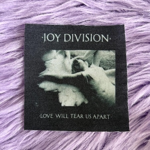 May include: A square black fabric patch featuring the white text JOY DIVISION at the top and LOVE WILL TEAR US APART at the bottom. The center displays a black and white graphic illustration of a figure lying down in a reclined position.