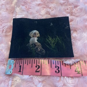 May include: A black and white patch with a woman in a white shirt and a brown jacket standing in a dark forest. The patch is approximately 3 inches wide.