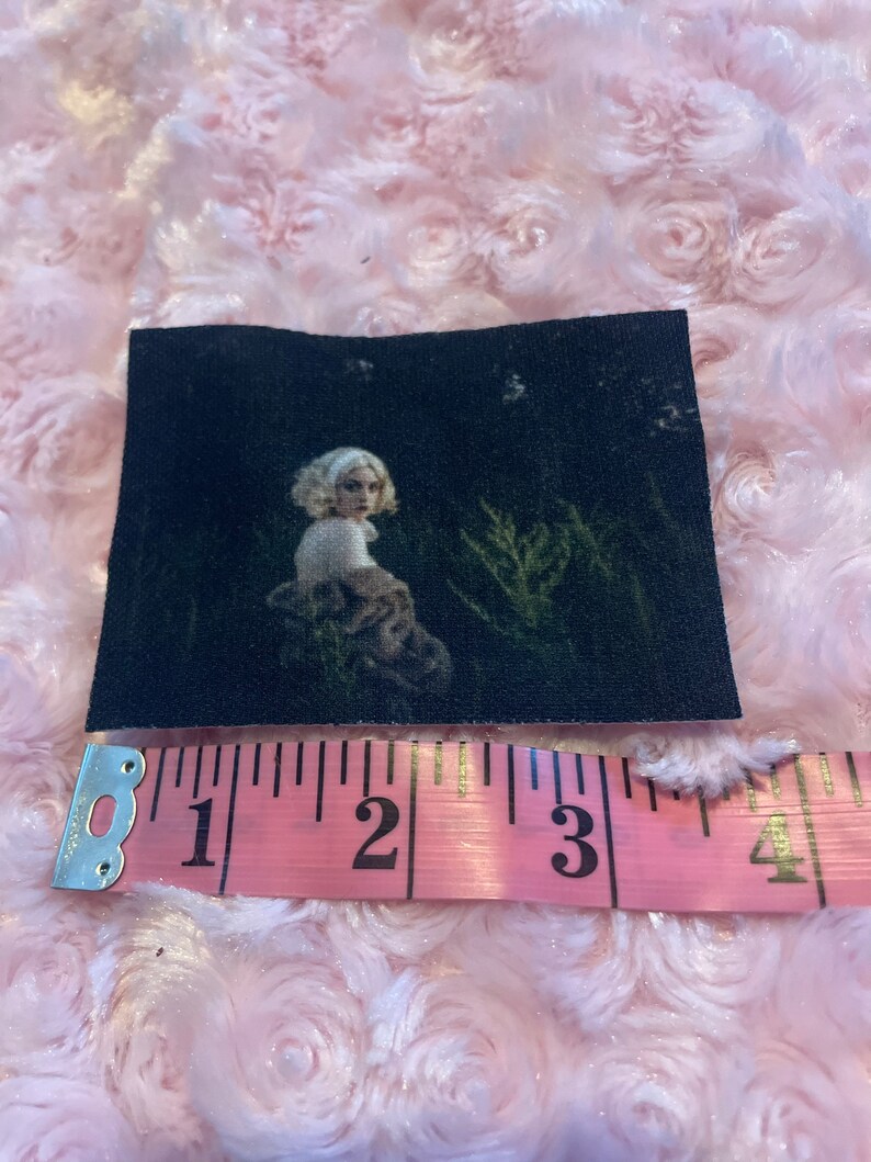 May include: A black and white patch with a woman in a white shirt and a brown jacket standing in a dark forest. The patch is approximately 3 inches wide.