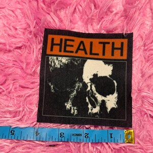 May include: A black patch with the word "HEALTH" in bold white letters on an orange background. Below the text is a black and white image of a skull. A blue measuring tape is placed below the patch.
