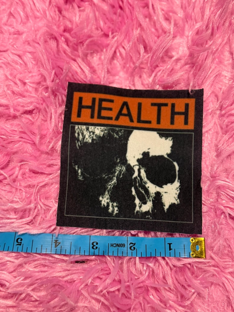 May include: A black patch with the word "HEALTH" in bold white letters on an orange background. Below the text is a black and white image of a skull. A blue measuring tape is placed below the patch.