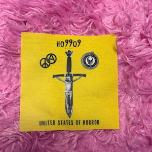 May include: A yellow patch with the text "H09909" and "UNITED STATES OF HORROR". The patch features a peace sign, a skull, and a crucified figure on a sword. The background is pink.