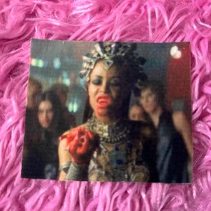 May include: A woman with dark hair and a jeweled headpiece holds a red heart in her hand. She is wearing a silver and black costume with a jeweled necklace. The background is a blurry city scene.