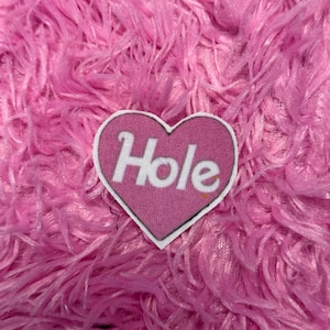 May include: A pink heart-shaped patch with the word "Hole" in white lettering. The patch is set against a background of fluffy, pink fabric.