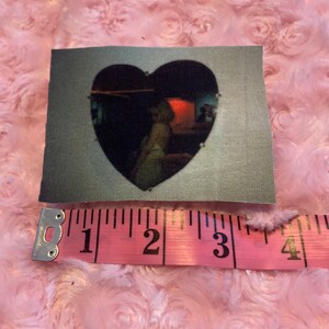 May include: A heart-shaped patch with a black and white image of a person inside a room with a red light. The patch is made of a gray fabric and measures approximately 2 inches in diameter.