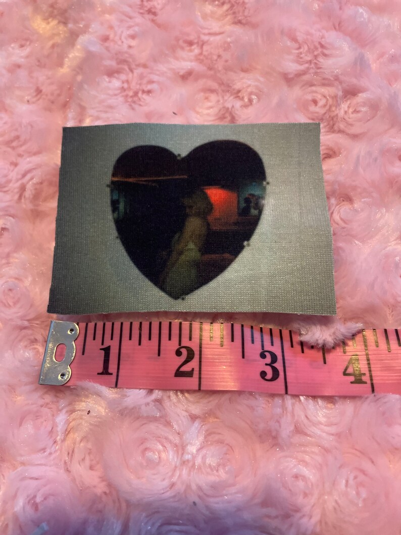 May include: A heart-shaped patch with a black and white image of a person inside a room with a red light. The patch is made of a gray fabric and measures approximately 2 inches in diameter.