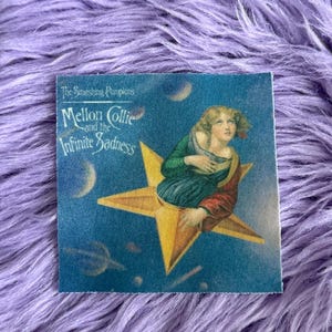 May include: A square fabric patch featuring the album art for The Smashing Pumpkins Mellon Collie and the Infinite Sadness. The design shows a person in a green and red robe sitting on a yellow star against a blue space background with moons.