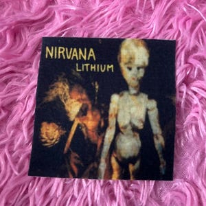 May include: A black and white iron-on patch with the band name "NIRVANA" and the song title "LITHIUM" printed in yellow. The patch features a black and white image of a person with long hair and a doll-like figure.