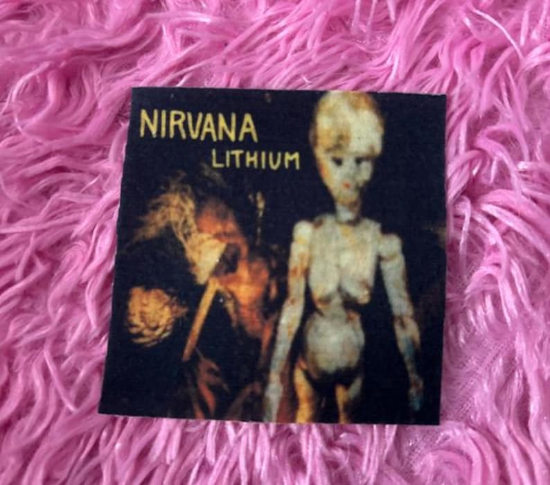 May include: A black and white iron-on patch with the band name "NIRVANA" and the song title "LITHIUM" printed in yellow. The patch features a black and white image of a person with long hair and a doll-like figure.