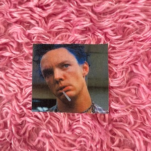May include: A close-up of a man with blue hair, wearing a plaid shirt and a necklace. He is holding a cigarette in his mouth and looking at the camera.