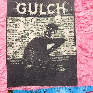 May include: A black and white patch with the word "GULCH" in a spiky font. The patch features a figure, mask designs, and the phrases "THE BODY IS A TEMPLE" and "THAT IS DAMNED TO DEMOLITION". The patch is approximately 10 cm wide.