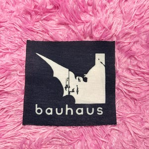 May include: A black square patch with the word "bauhaus" in white, beneath a white graphic design. The graphic appears to be a stylized bat or bird shape. The patch is set against a pink, fluffy background.