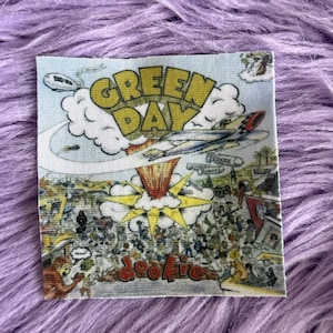 May include: A square fabric patch featuring the Green Day Dookie album cover art. The design includes a yellow logo, a central explosion, a flying airplane, and a detailed chaotic scene with various figures and buildings in muted colors.