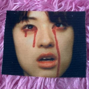 May include: A rectangular patch featuring a close-up of a person's face with red streaks resembling tears flowing from the eyes. The person has dark hair, pale skin, and red lipstick. The background is a textured black.