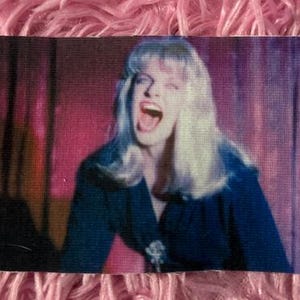 May include: A woman with long blonde hair is singing with her mouth open wide. She is wearing a dark blue shirt and is in a dimly lit room with purple and pink lighting.