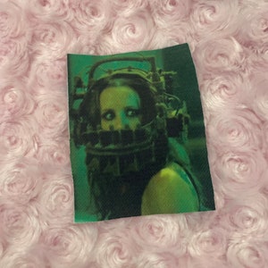 May include: A close-up of a woman's face wearing a metal cage over her head. The cage is rusty and has sharp edges. The woman's eyes are wide with fear. The background is dark and blurry.