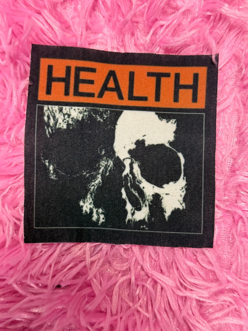 May include: A black patch with the word "HEALTH" in orange block letters above a white skull graphic on a black background. The patch is set against a vibrant pink, fluffy background.