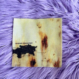 May include: A square fabric patch featuring the Nine Inch Nails The Downward Spiral album cover art. The design displays a pale yellow background with brown rust-like stains and a prominent black abstract shape on the left side with text.