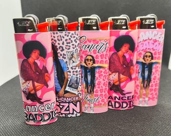 May include: Five rectangular lighters featuring pink and black graphic designs. Each lighter displays images of individuals with dark hair, leopard print patterns, and pink swirls. The text "Cancer Baddie," "Cancer SZN," and "Cancer Season" is printed on the lighters in bold, stylized lettering.