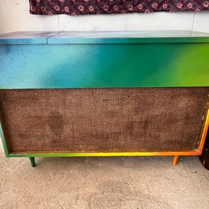 May include: A vintage stereo cabinet with a gradient paint job in shades of blue, green, and yellow. The cabinet has a woven speaker cover and sits on short, angled legs. The top of the cabinet has a hinged lid.