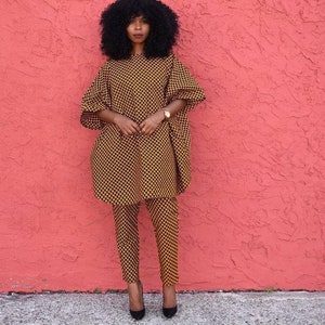 May include: A woman wearing a yellow and black patterned trouser suit with a long-sleeved top and trousers. She is standing in front of a pink wall.