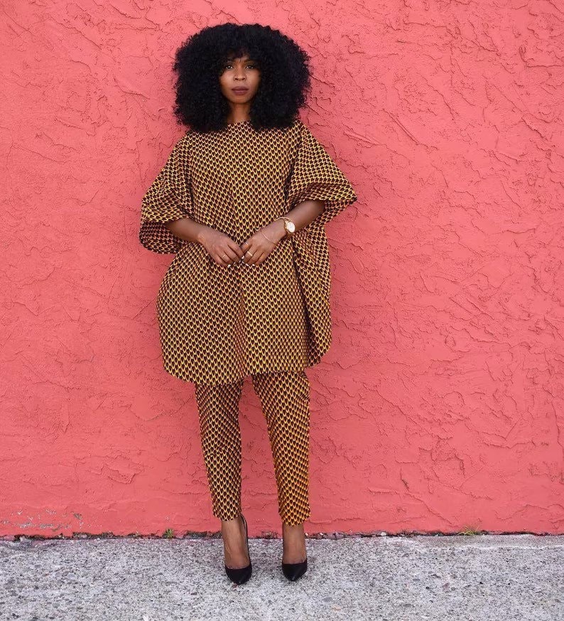 May include: A woman wearing a yellow and black patterned trouser suit with a long-sleeved top and trousers. She is standing in front of a pink wall.