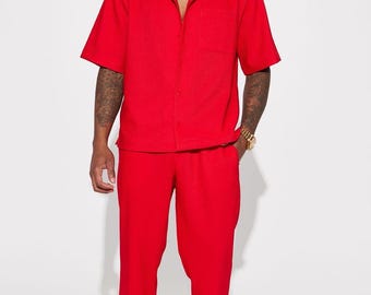 May include: A red two-piece outfit consisting of a short-sleeved button-down shirt with a chest pocket and matching pants. The outfit is made of a textured fabric. The model is wearing a gold chain and watch.