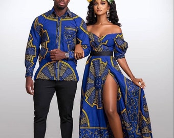 Op de afbeelding: Een stel draagt ​​bijpassende blauwe en gouden shirts met patronen. De vrouw draagt ​​een lange jurk met een hoge split en een bloemenkrans. Ze staan ​​voor een witte achtergrond.