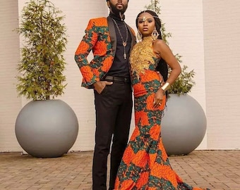 May include: A couple wearing matching orange and green patterned outfits. The man is wearing a blazer and black pants. The woman is wearing a sleeveless dress with a gold bodice. They are standing in front of a white wall with potted plants.