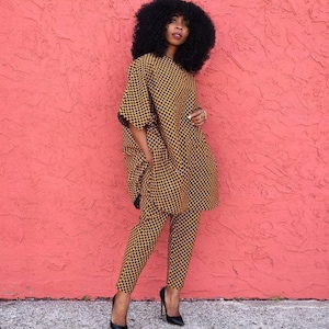 May include: A woman wearing a yellow and black patterned trouser suit with a long-sleeved top and trousers. She is standing in front of a pink wall.