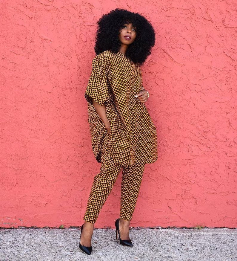 May include: A woman wearing a yellow and black patterned trouser suit with a long-sleeved top and trousers. She is standing in front of a pink wall.